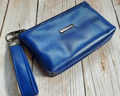 side view of a blue marbled faux leather makeup bag with gunmetal hardware and a gunmetal Bass Creations logo. It also includes a wristlet strap for easy carrying.