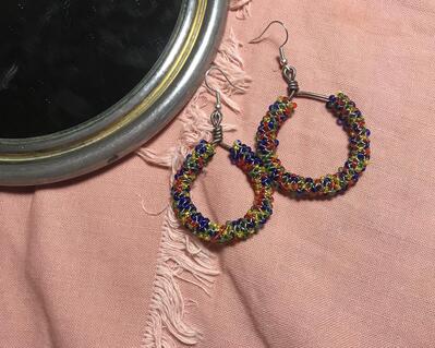 small rainbow seed bead peyote stitch hoop earrings. beaded tube is on a mild steel hoop frame with silver-plated earring hooks. photographed on a pink fabric background.
