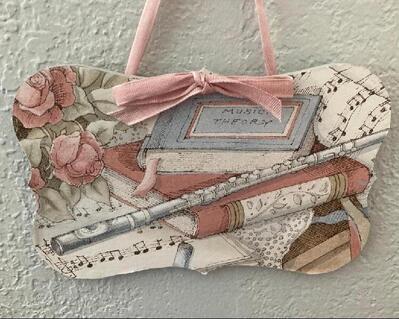 Decorative music plaque in pink, brown, white, and black. plaque has rounded dimensions and highlights the covering.. It is more of an item from the victorian period.  Soft colors with multiple instruments and music text.  The flute, music manuscript, and literature books enhance the entire plaque.
