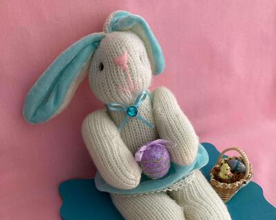 A white sock Easter bimmy seated on a turqouise decorative plaque.  The bunny is wearing a skirt with strapes made in turquoise velour and ears are same color.  Her bow has an aqua crystal.  A small basket has eggs and a chicken as does the decorative plaque.
