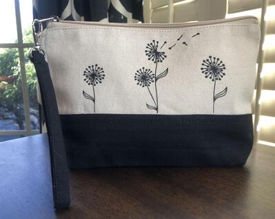 9 inch wristlet with black canvas on the bottom and cream canvas on the top, featuring three black vinyl dainty dandelions on each side, a black removable strap, soft cream interior lining and a flat bottom for extra capacity