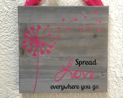 10 inch square gray-washed wood sign featuring a pink dandelion and the words spread love everywhere you go, the word love is in pink and the remaining words in black, a pink ribbon is attached for hanging, a sawtooth picture hanger is also attached to the back