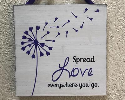 white-washed 10 inch square sign featuring a purple dandelion and the words spread love everywhere you go, the word love is in purple, the remaining words in black, a purple ribbon is attached to the back for hanging