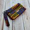 Side view of a golden yellow, green and blue Kente printed makeup pouch with 2 gunmetal toned zipper pockets and a gunmetal toned Bass Creations logo.