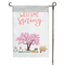 White and pink garden flag with flowering tree, bicycle, wheel barrow, and rain boots.
