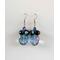 This pair of dangle earrings features a large blue faceted glass bead that switches to purple in the light. There are 4 black round beads seated on top of that with an AB finish on them, bringing out purples, blues, greens, and yellows. Then another purple bead on top with an AB finish and they are connected to a silver earring hook.