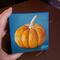 Small square painting of little orange pumpkin with teal blue background.