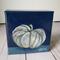 Photo of small square canvas painting of oddly shaped light blue pumpkin with a dark blue background