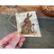 A handmade gift tag. It is a piece of real birch bark with a woodland rabbit wood burned onto the bark. A jute cord is tied in the corner. 