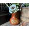 This shows a carved gourd vase with faux flowers. The vase is decorating a fireplace hearth. It sits next to an old wood barrel. 