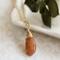 Sunstone necklace, orange briolette in 14K gold, on white dish with green leaves.
