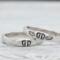 Two sterling silver rings with hand-stamped baby feet and wings, placed on a white wooden surface with delicate white flowers.
