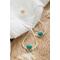 "Pair of silver drop earrings with turquoise gemstones, resting on a white fabric with dried flowers on a wooden surface."