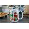 white mug with 4 different Christmas themed  iced coffee cups printed around it. One with candy canes, one with Poinsette, one with a snowman, and one with peppermint candies. Presents, gingerbread man and snowman cookie sit in between the cups.