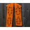 Orange scarf with leaves along the length. It is on a hanger in front of a wooden fence.