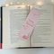 The romantic bookmark is sitting in the middle of an open book with fairy lights around the book on a white table