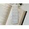 the laminated bookmark is on top of an open book with a pair of glasses above the book and fairy lights on the right side on a white table