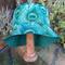Front view of bucket hat on stand. All over print features drawn peacock feathers on light blue background. Part of the reverse side of hat is visible to show the coordinating blue-green.