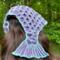 A woman with dark brown hair wearing a mermaid tail shaped crochet hair bandana that has medium purple scales with light blue outlines.