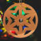 A handmade wooden Christmas tree ornament, circular in shape, crafted from light brown wood with a natural grain pattern. It features an intricate, symmetrical cutout design resembling a star or snowflake, with multiple pointed shapes radiating from a central hole. The ornament is attached to a small metal hook for hanging, showcasing its rustic and festive craftsmanship.