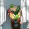 Hummingbird stained glass night light- made of green white and red glass, background is of light blue glass with a pink flower and lampworked green leaf. Show with light on.