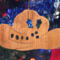 Handmade wooden Christmas tree ornament shaped like a cowboy hat, featuring a sprig of holly tucked into the hat band, star-shaped leaf cutouts, and round berry accents.