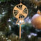 A handcrafted wooden Christmas tree ornament shaped like a lollipop, featuring intricate cut-out spiral patterns on the circular top and a simple stick base. The ornament is light brown with a smooth finish.