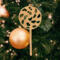 A handmade Christmas tree ornament crafted from light brown maple hardwood plywood, shaped like a lollipop with a round head featuring intricate spiral cutouts and a long stick handle, suspended from a small metal hook.