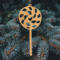 A handmade Christmas tree ornament crafted from light brown maple hardwood plywood, shaped like a lollipop with a round head featuring intricate spiral cutouts and a long stick handle, suspended from a small metal hook.
