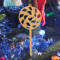 A handmade Christmas tree ornament crafted from light brown maple hardwood plywood, shaped like a lollipop with a round head featuring intricate spiral cutouts and a long stick handle, suspended from a small metal hook.