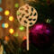 A handmade Christmas tree ornament crafted from light brown maple hardwood plywood, shaped like a lollipop with a round head featuring intricate spiral cutouts and a long stick handle, suspended from a small metal hook.