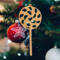 A handmade Christmas tree ornament crafted from light brown maple hardwood plywood, shaped like a lollipop with a round head featuring intricate spiral cutouts and a long stick handle, suspended from a small metal hook.