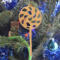 A handmade Christmas tree ornament crafted from light brown maple hardwood plywood, shaped like a lollipop with a round head featuring intricate spiral cutouts and a long stick handle, suspended from a small metal hook.