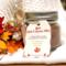 Handmade hot cocoa mix jar with wooden spoon and red-gold ribbon, displayed on a white knit blanket with autumn leaves.