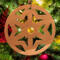 A handmade wooden Christmas tree ornament, circular in shape, crafted from light brown wood with a natural grain pattern. It features an intricate, symmetrical cutout design resembling a star or snowflake, with multiple pointed shapes radiating from a central hole. The ornament is attached to a small metal hook for hanging, showcasing its rustic and festive craftsmanship.