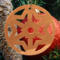 A handmade wooden Christmas tree ornament, circular in shape, crafted from light brown wood with a natural grain pattern. It features an intricate, symmetrical cutout design resembling a star or snowflake, with multiple pointed shapes radiating from a central hole. The ornament is attached to a small metal hook for hanging, showcasing its rustic and festive craftsmanship.