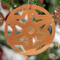 A handmade wooden Christmas tree ornament, circular in shape, crafted from light brown wood with a natural grain pattern. It features an intricate, symmetrical cutout design resembling a star or snowflake, with multiple pointed shapes radiating from a central hole. The ornament is attached to a small metal hook for hanging, showcasing its rustic and festive craftsmanship.