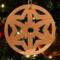 A handmade wooden Christmas tree ornament, circular in shape, crafted from light brown wood with a natural grain pattern. It features an intricate, symmetrical cutout design resembling a star or snowflake, with multiple pointed shapes radiating from a central hole. The ornament is attached to a small metal hook for hanging, showcasing its rustic and festive craftsmanship.