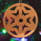 A handmade wooden Christmas tree ornament, circular in shape, crafted from light brown wood with a natural grain pattern. It features an intricate, symmetrical cutout design resembling a star or snowflake, with multiple pointed shapes radiating from a central hole. The ornament is attached to a small metal hook for hanging, showcasing its rustic and festive craftsmanship.