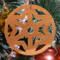 A handmade wooden Christmas tree ornament, circular in shape, crafted from light brown wood with a natural grain pattern. It features an intricate, symmetrical cutout design resembling a star or snowflake, with multiple pointed shapes radiating from a central hole. The ornament is attached to a small metal hook for hanging, showcasing its rustic and festive craftsmanship.