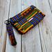 Side view of a golden yellow, green and blue Kente printed makeup pouch with 2 gunmetal toned zipper pockets and a gunmetal toned Bass Creations logo.
