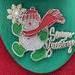 Decorative sign of snowman and Season Greetings.  Plastic.  Santa is also holding a candy cane, gold pinecone on hat, and greenery on opposite side of his body. Green plaque.