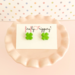 Pair of green four leaf clover stud earrings on a fireflyFrippery branded jewelry card resting on a miniature, soft pink cupcake stand in front of a pastel polka-dot background