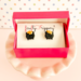 Pair of pot of gold stud earrings on a fireflyFrippery branded jewelry card and nestled in a hot pink gift box with lid, resting on a miniature, soft pink cupcake stand in front of a pastel polka-dot background