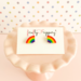 Pair of half rainbow stud earrings on a fireflyFrippery branded jewelry card resting on a miniature, soft pink cupcake stand in front of a pastel polka-dot background