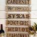 A wine sign with Cabernet, Prosecco, Syrah, Pinot Gris, Merlot, and Chardonnay wood-burned onto boards and then tied together with twine. 