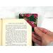 This shows a pink flower fabric corner bookmark with a dark emerald green fabric applique heart on the front.  You can see the same green fabric is used for the base of the corner bookmark too.  It's showing the bookmark being slid over the corner of a page of a book.