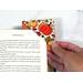 A fun fall fabric design was used to make a corner bookmark with leaves, pumpkins, etc.  The bottom of the bookmark is orange and the picture depicts it being slid over a page in a book to demonstrate how to use this handmade bookmark corner.