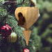 The wooden miniature Birdhouse Ornament is shaped like a heart. A small, dark-reddish-brown bird is perched on the front of the birdhouse. The heart-shaped structure has a pointed bottom that tapers down to a fine point. The wood is light beige.