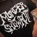 Black t-shirt on bigger, tattooed model holding her hand up presentation style with just the logo/torso showing.  The shirt image is a brutal font meshed together reading Murder She Wrote in the style of a Metal band logo.